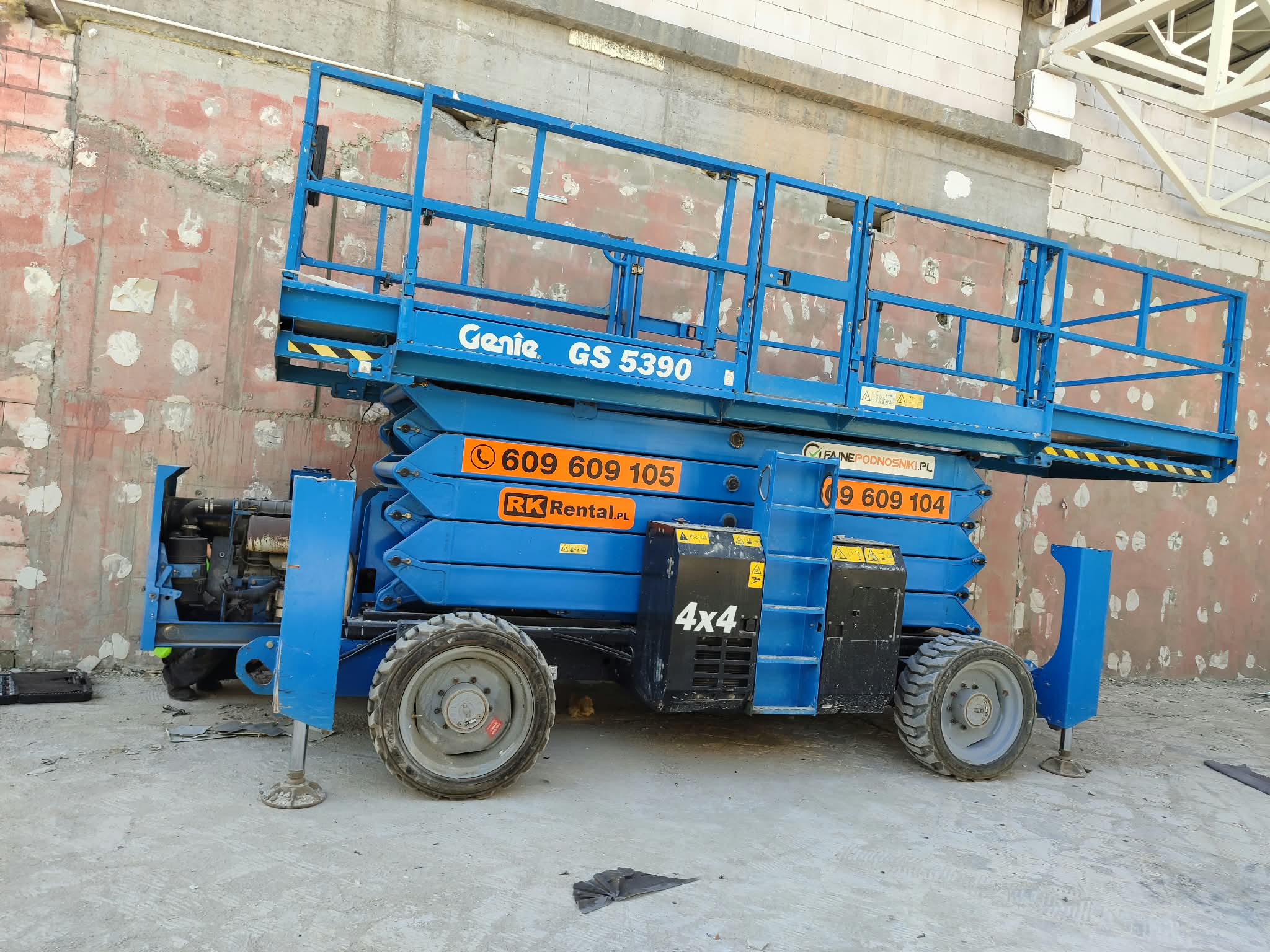 Genie GS-5390 RT rough terrain scissor lift (18 m, 4×4) from RK Rental on outriggers next to the wall of a building under construction in Gostynin