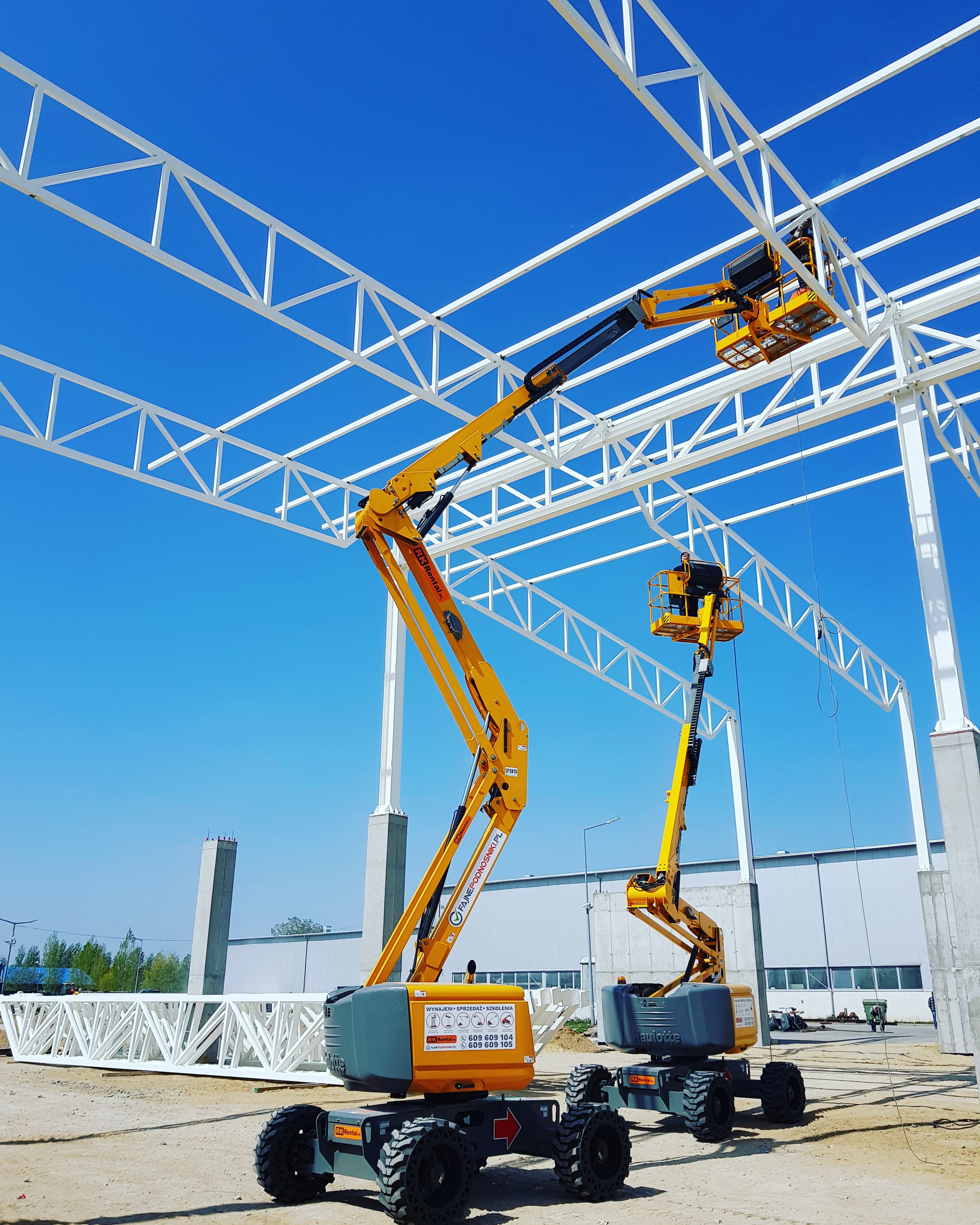 Dwa terenowe podnośniki przegubowe Haulotte HA16 RTJ (16 m, 4×4) RK Rental przy montażu konstrukcji stalowej hali w Poznaniu